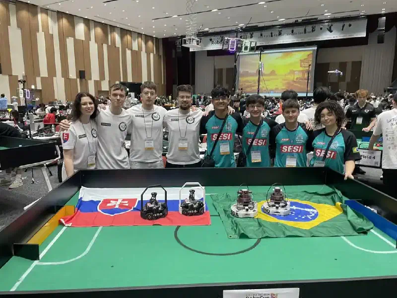 Team XLC – WYLDFYRE and the Brazilian team standing behind a RoboCup soccer field, with both national flags and both teams' robots placed on the green playing surface