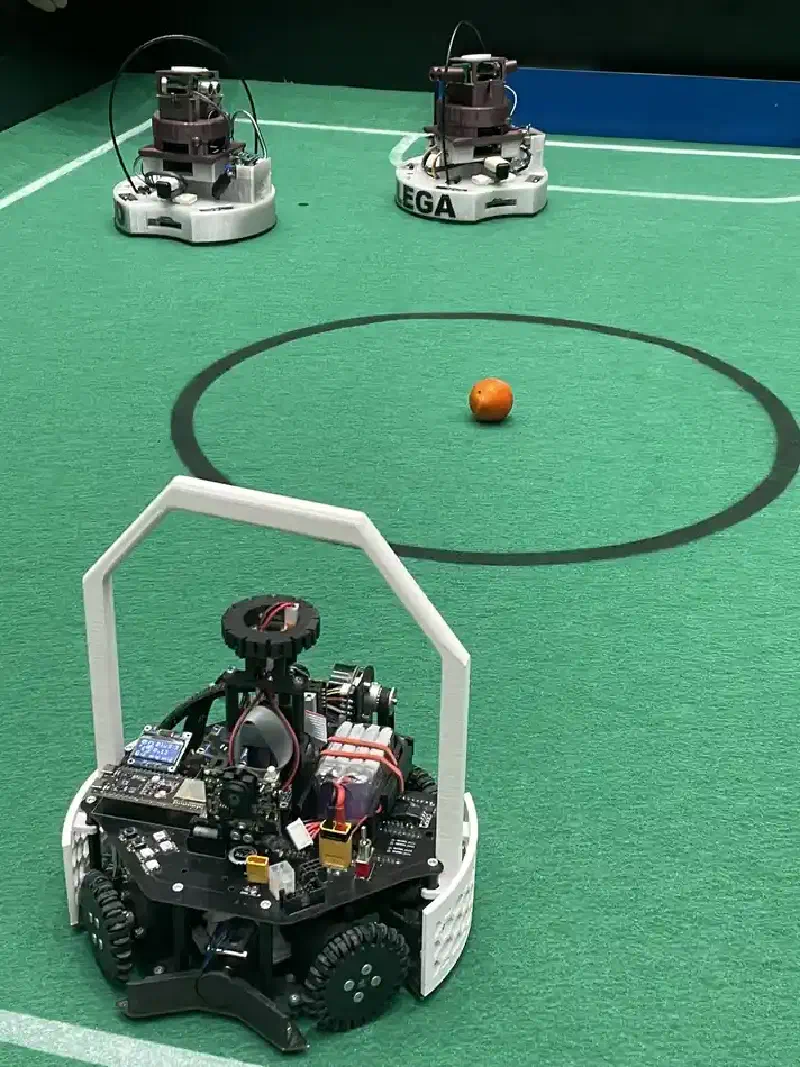 A RoboCup Soccer match in progress: our black robot in the foreground on the green field with the orange infrared ball nearby, two opponent robots in the background