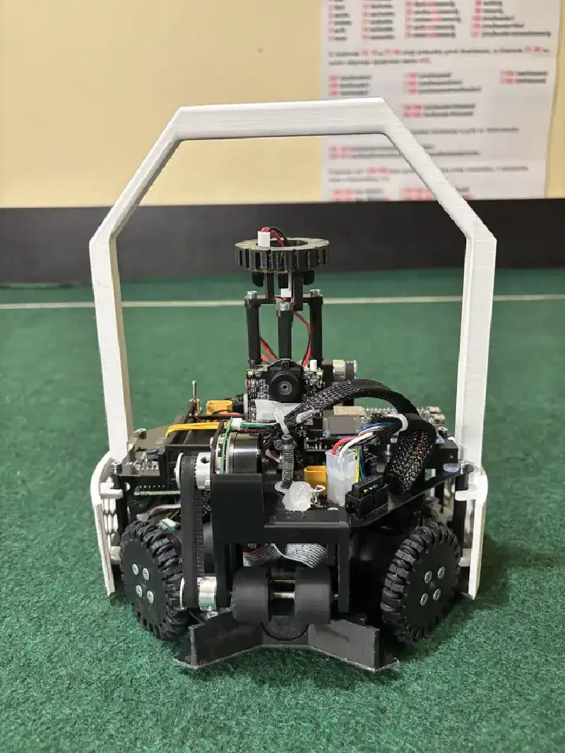 Front view of the robot on the green field: the dribbler roller and its belt drive sit low at the front between two omni wheels, with the Pixy camera and the electronics stack above