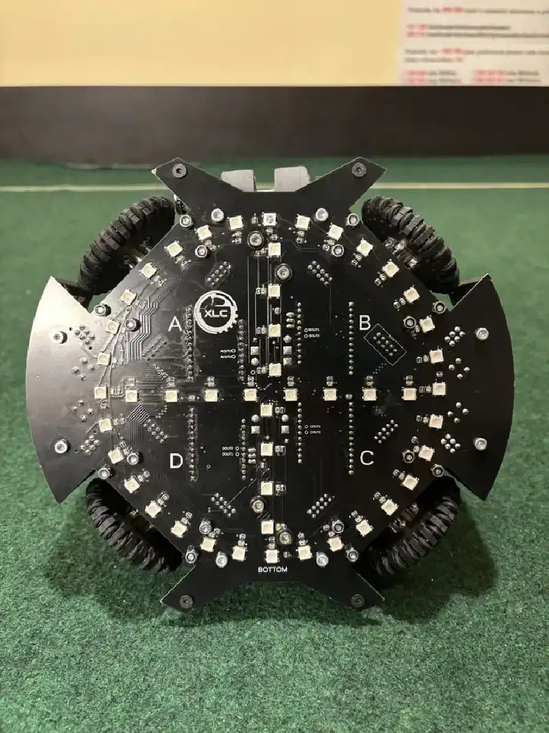 The underside of the robot: a black four-lobed circuit board with a ring of surface-mount RGB LEDs and phototransistors around the perimeter, four omni wheels in cut-outs labelled A, B, C and D, and the XLC logo at the centre