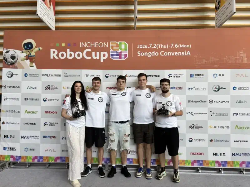 The four members of team XLC – WYLDFYRE and their mentor standing in front of the Incheon RoboCup 2026 sponsor backdrop, two of them holding their robots